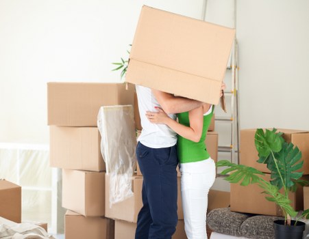 Two individuals are engaged in a house move scene within a well-lit room, possibly a living area or hallway, where they are lifting a medium-sized cardboard box together. The person on the left, wearing a white t-shirt and blue jeans, holds the box from the bottom, while the person on the right, dressed in a bright green top and white trousers, supports the box from the side. In the background, several other cardboard boxes of various sizes are stacked, with some wrapped in white packing paper, indicating they are prepared for transport. A potted plant with large green leaves is positioned on a small table or stand to the right, adding a natural element to the scene. The room has neutral-colored walls and the floor appears to be a light wood or laminate, suggesting a modern residential property in Belsize Park, London, being prepared for a house removal with Belsize Park Man with Van. Bright, natural lighting enhances the clarity of the scene and highlights the textures of the cardboard, clothing, and houseplants.
