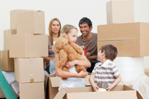 In a well-lit indoor setting, a family is surrounded by numerous brown cardboard moving boxes, some stacked on top of each other, indicating an ongoing house move. An adult woman with long blonde hair stands in the background, while an adult man with dark hair and a beard stands nearby, both smiling. In the foreground, a young girl with blonde hair, dressed in a beige fluffy coat, is holding a small teddy bear and facing a young boy with short brown hair, who is wearing a striped polo shirt. The boy is standing in an open cardboard box, possibly being used for packing or unpacking. The scene captures a moment of interaction and preparation, with household items and boxes arranged around them, suggesting a house relocation process in progress. The environment appears to be a bright, neutral-colored room, fitting for residential house removals in the Belsize Park area. This image exemplifies the typical activity of a family packing or preparing for a move, illustrating the kind of service offered by Belsize Park Man with Van in north London.