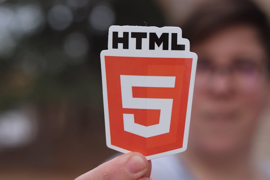 A person is holding a small, laminated sticker with the HTML5 logo, featuring a red shield shape with a white number 5 and black text reading 'HTML'. The individual is partially visible, with blurred 