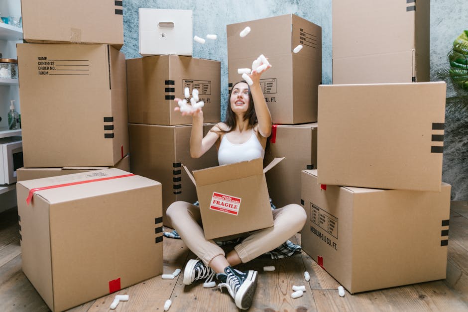 A young woman with curly hair is standing in the doorway of a brick building, carrying two cardboard boxes, one in each arm. She is wearing a white t-shirt, blue jeans, and sneakers, with a red and bl